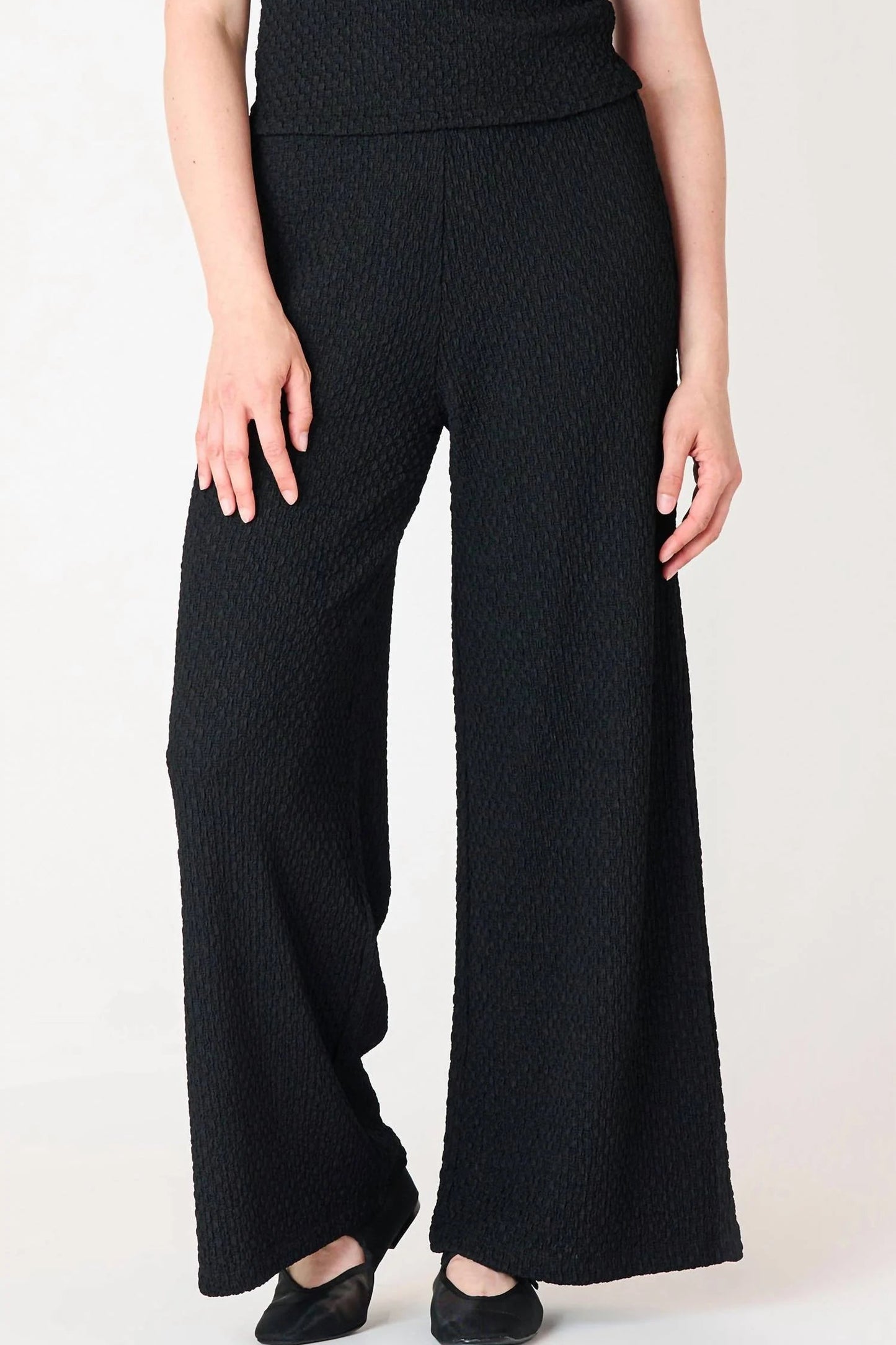 A model wearing black wide-leg knit pants with a textured waffle pattern, paired with a matching black top and black flats, shown against a plain white studio background.