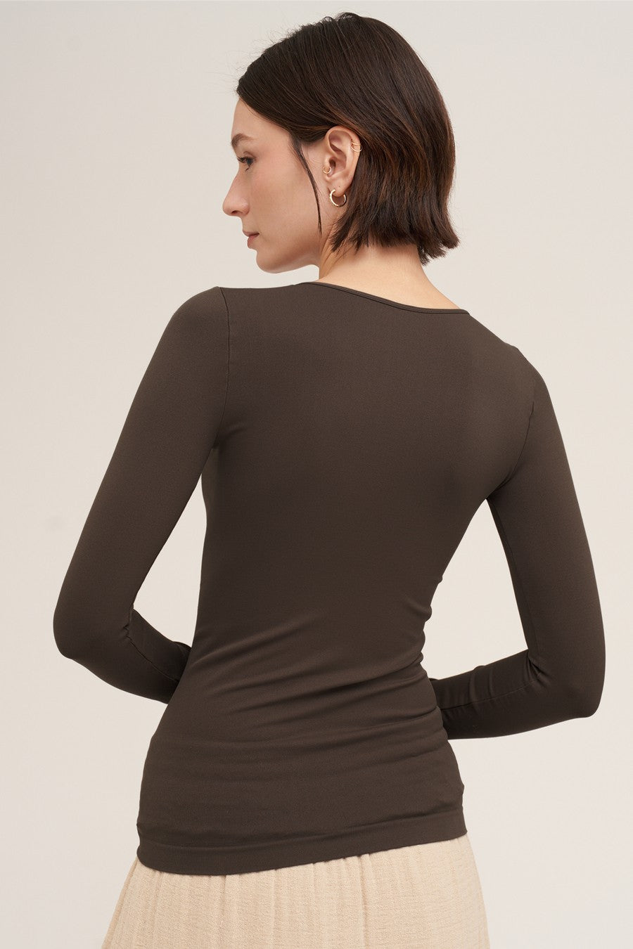 A woman shown from the back wearing a fitted long-sleeve top in a dark brown color, paired with a light beige skirt, against a plain light beige studio background.