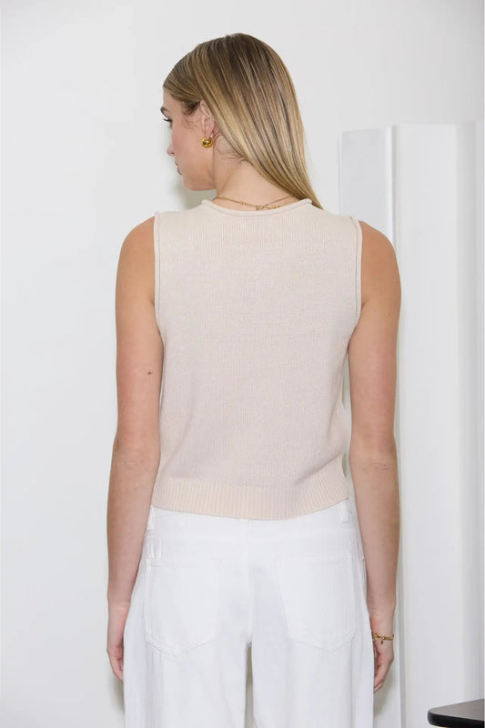 Woman wearing a beige sleeveless top and white pants against a white background