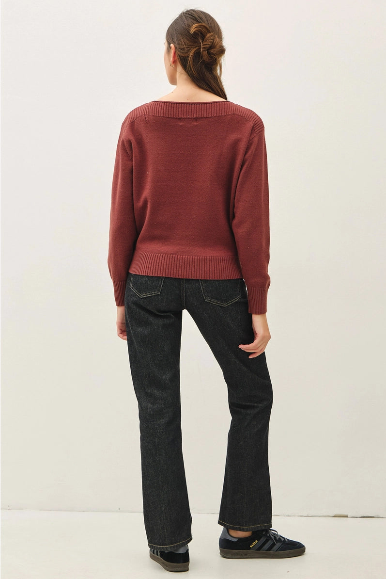 A woman viewed from behind wearing a rust-red knit sweater, dark wash jeans, and black sneakers against a plain off-white studio background.