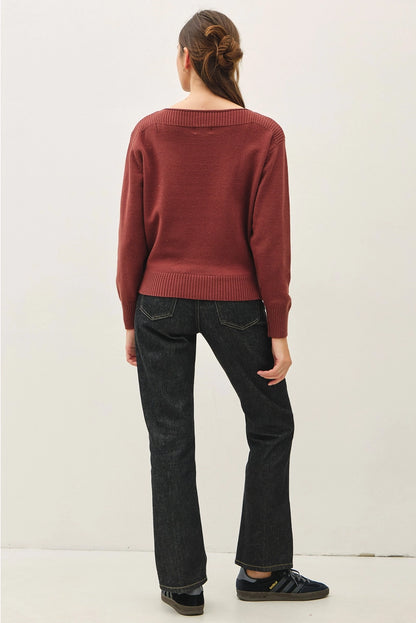 A woman viewed from behind wearing a rust-red knit sweater, dark wash jeans, and black sneakers against a plain off-white studio background.