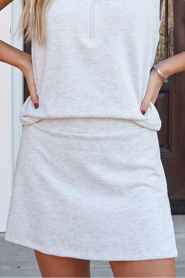 A woman wearing a light heather gray sleeveless collared pullover dress with a short front zipper and side pockets, standing outdoors in front of a doorway.