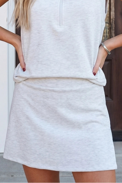 A woman wearing a light heather gray sleeveless collared pullover dress with a short front zipper and side pockets, standing outdoors in front of a doorway.