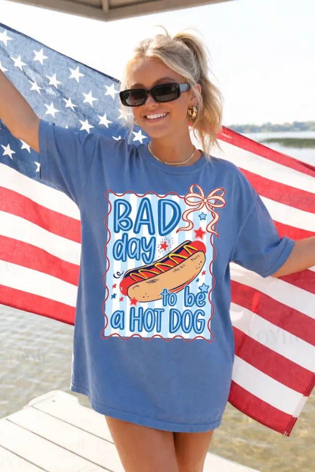 Woman wearing a blue t-shirt with a hot dog graphic and text, standing in front of an American flag by a body of water.