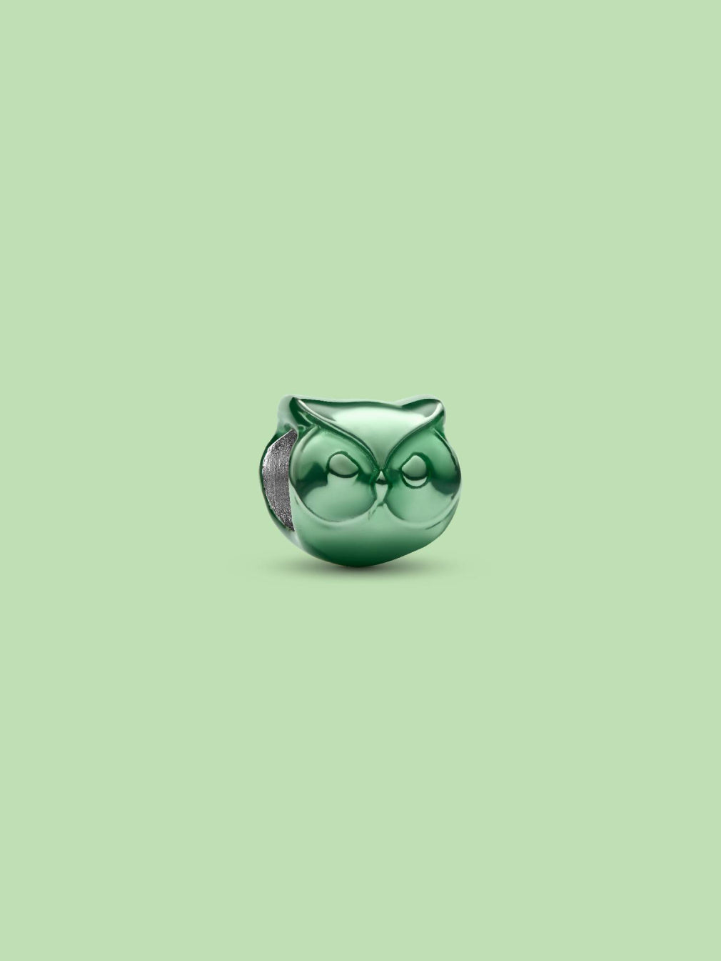 A small glossy green owl-shaped charm or bead with a silver-toned side detail, centered on a solid pale green background.
