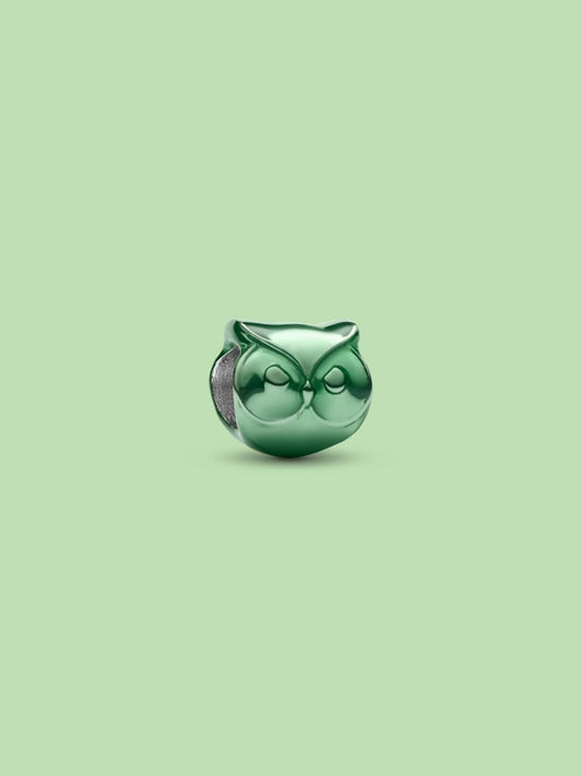 A small glossy green owl-shaped charm or bead with a silver-toned side detail, centered on a solid pale green background.