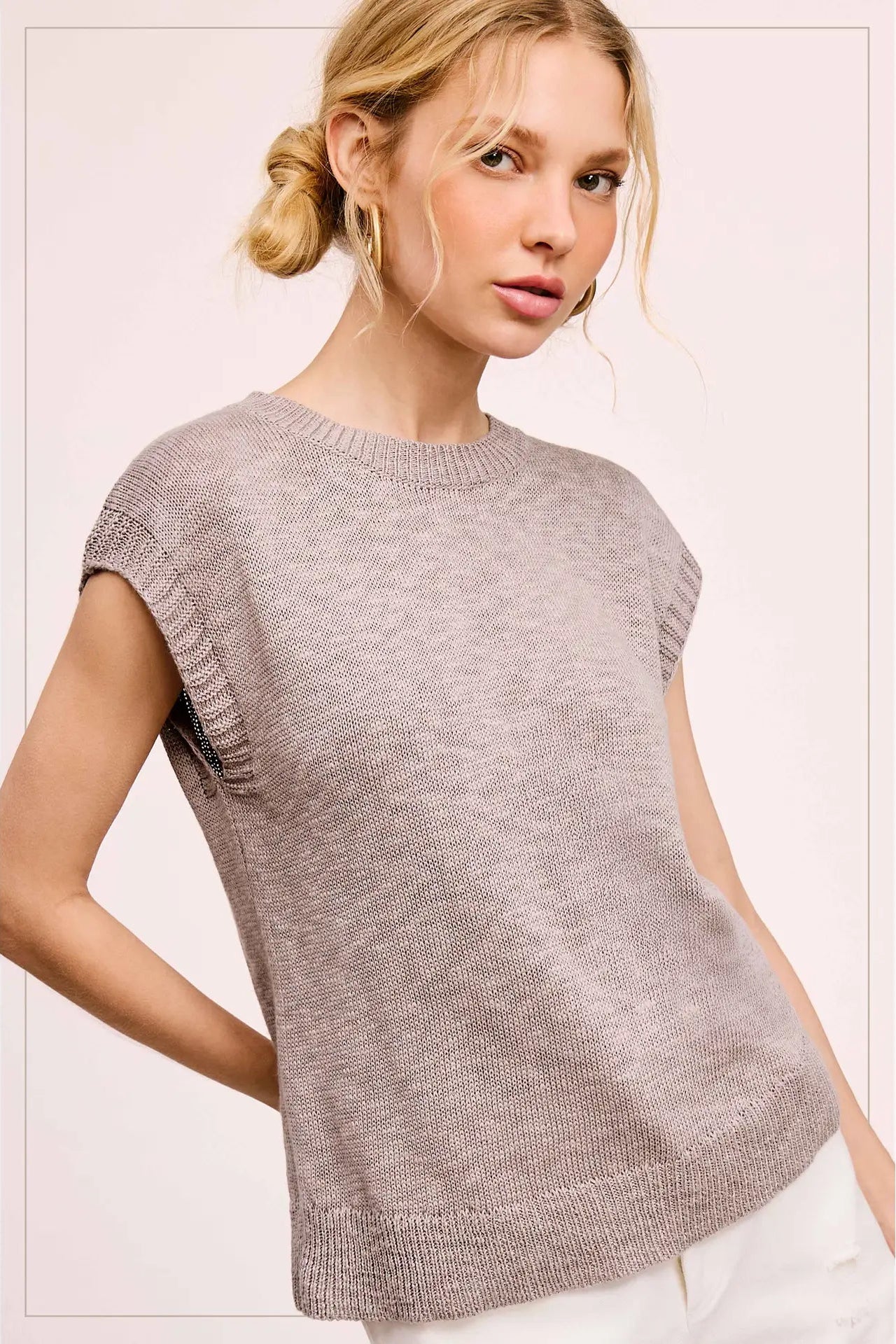 A woman wearing a light heather gray knit sweater with cap sleeves and a crew neck, paired with white pants, set against a pale pink studio background.