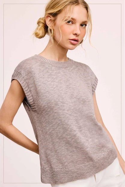 A woman wearing a light heather gray knit sweater with cap sleeves and a crew neck, paired with white pants, set against a pale pink studio background.