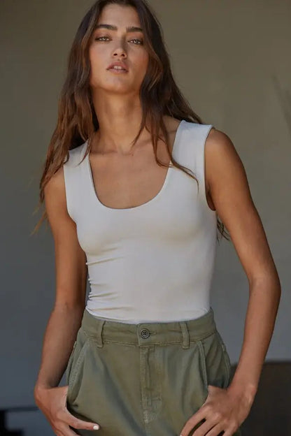 A woman wearing a fitted white scoop-neck tank top and olive green pants, standing against a neutral indoor background.