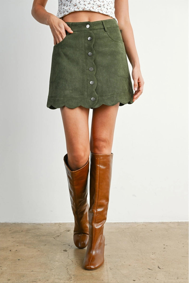 An olive green corduroy mini skirt with a scalloped hem and front button closure, worn by a model with brown knee-high leather boots against a neutral studio background.
