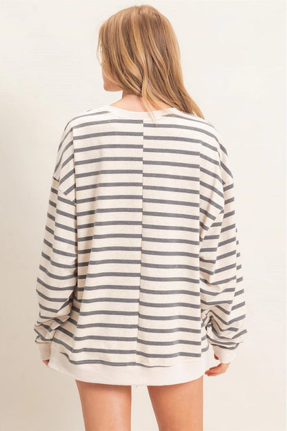 A woman viewed from the back wearing an oversized long-sleeve top with horizontal gray stripes on a cream base, set against a plain light beige studio background.