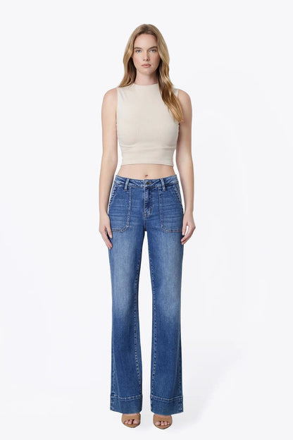 A woman modeling a pair of high-waisted, wide-leg blue jeans with a beige sleeveless crop top, standing against a plain white studio background.