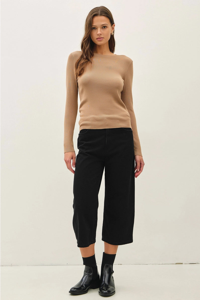 A woman modeling a beige long-sleeve knit top and black cropped pants with black socks and black boots, standing against a plain off-white studio background.