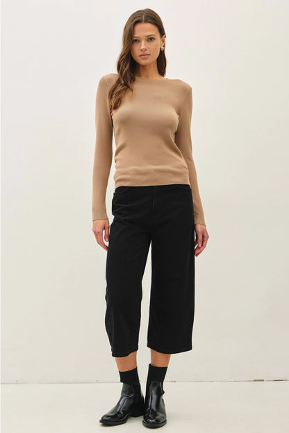 A woman modeling a beige long-sleeve knit top and black cropped pants with black socks and black boots, standing against a plain off-white studio background.