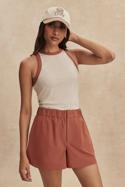 Woman wearing a white tank top, rust-colored shorts, and a white cap with a logo on a beige background