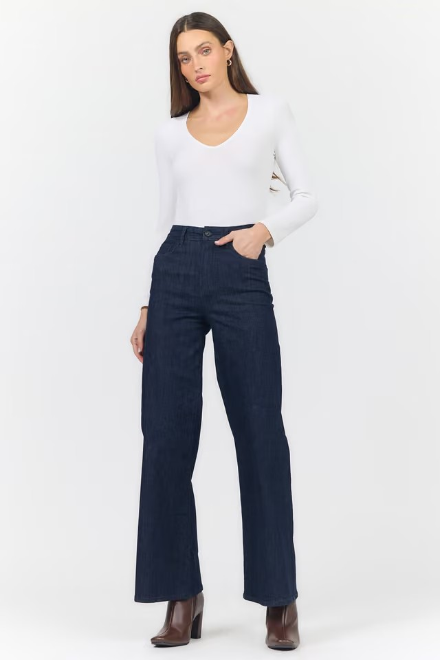 A woman modeling a pair of dark navy blue wide-leg jeans with a white long-sleeve top and brown ankle boots, standing against a plain white studio background.