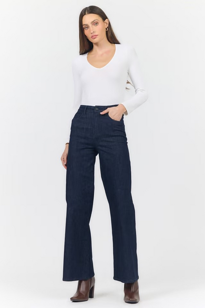 A woman modeling a pair of dark navy blue wide-leg jeans with a white long-sleeve top and brown ankle boots, standing against a plain white studio background.