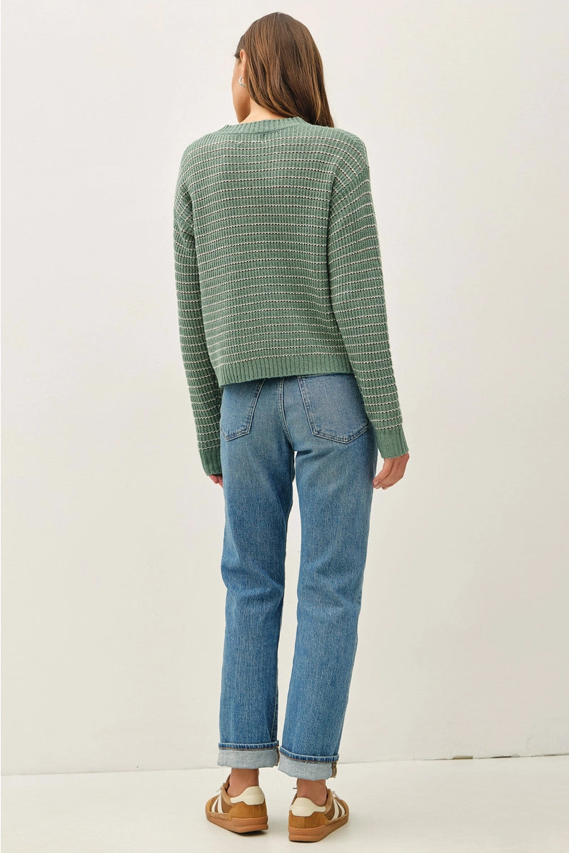 A woman shown from the back wearing a green and white striped knit sweater, blue jeans, and brown shoes, standing against a plain white studio background.
