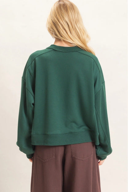 A woman viewed from behind wearing a dark green cropped sweatshirt and brown wide-leg pants against a plain off-white background.