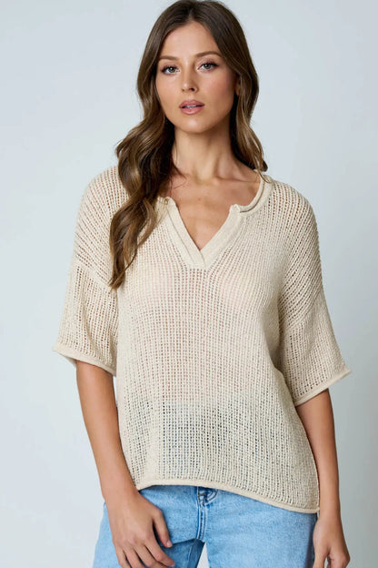 A woman wearing a beige open-knit short-sleeve V-neck top and light blue jeans, standing against a light gray studio background.