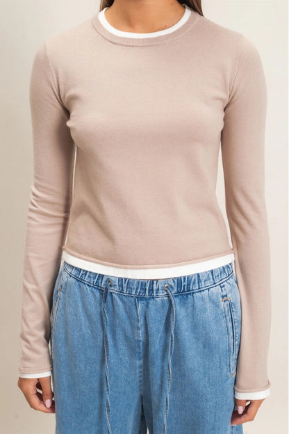 A woman wearing a fitted long-sleeve knit top in a light warm beige or blush tone with white contrast trim at the crew neckline, hem, and cuffs, paired with blue jeans against a plain light beige studio background.