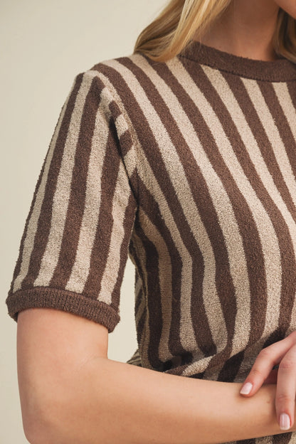 Close-up of a woman wearing a short-sleeve knit top with vertical brown and beige stripes, cropped from the neck to the forearm against a plain light beige background.