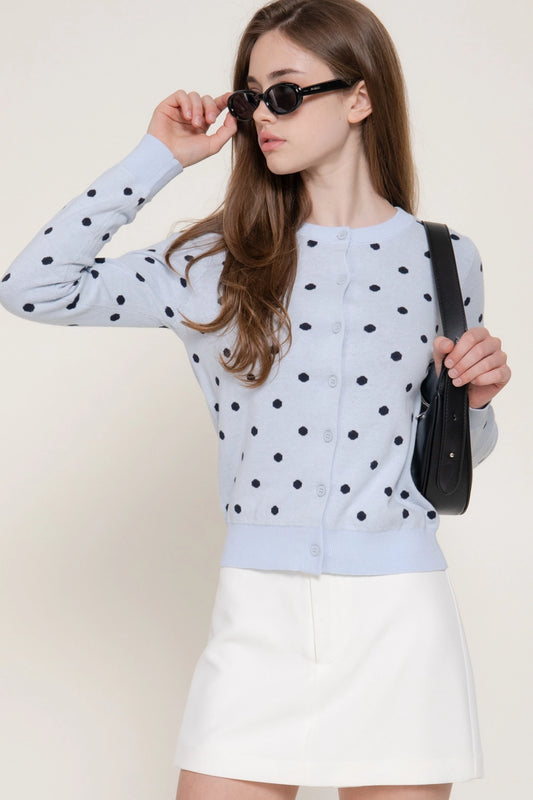 A woman wearing a light blue long-sleeve cardigan with black polka dots, a white mini skirt, and black sunglasses, holding a black shoulder bag against a plain beige background.
