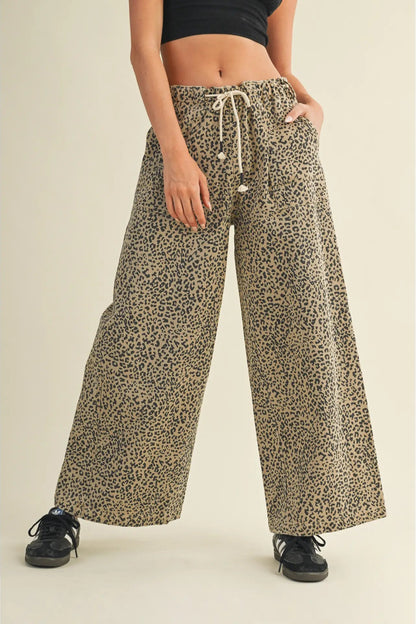 A person wearing loose, wide-leg leopard-print pants with a cream drawstring waist, paired with a black crop top and black sneakers, against a plain beige studio background.