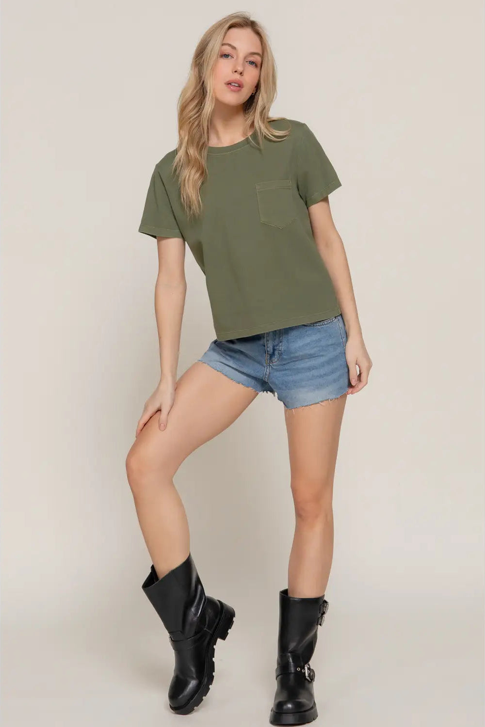 Woman wearing an olive green t-shirt, denim shorts, and black boots on a plain background