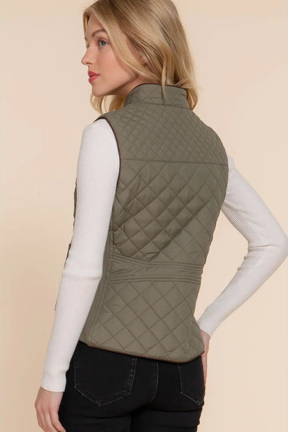 A woman shown from the back wearing an olive green quilted sleeveless puffer vest over a white ribbed long-sleeve top and black jeans, set against a solid beige studio background.
