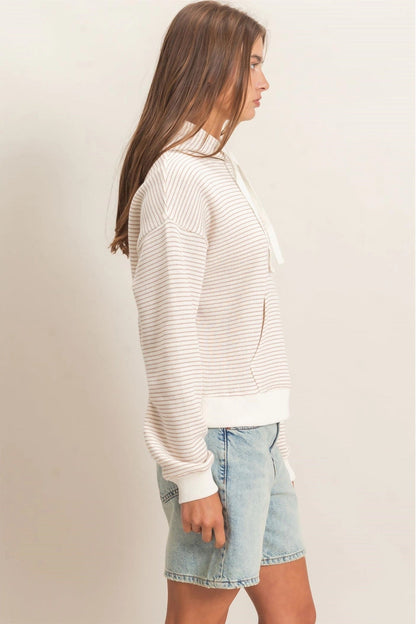 A woman shown in side profile wearing a cream and white horizontally striped knit hoodie with a drawstring, paired with light-wash denim shorts, against a plain beige studio background.