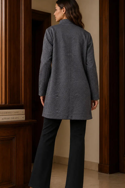 A woman shown from the back wearing a textured charcoal gray longline coat and black wide-leg trousers, standing in a room with dark wood paneling.