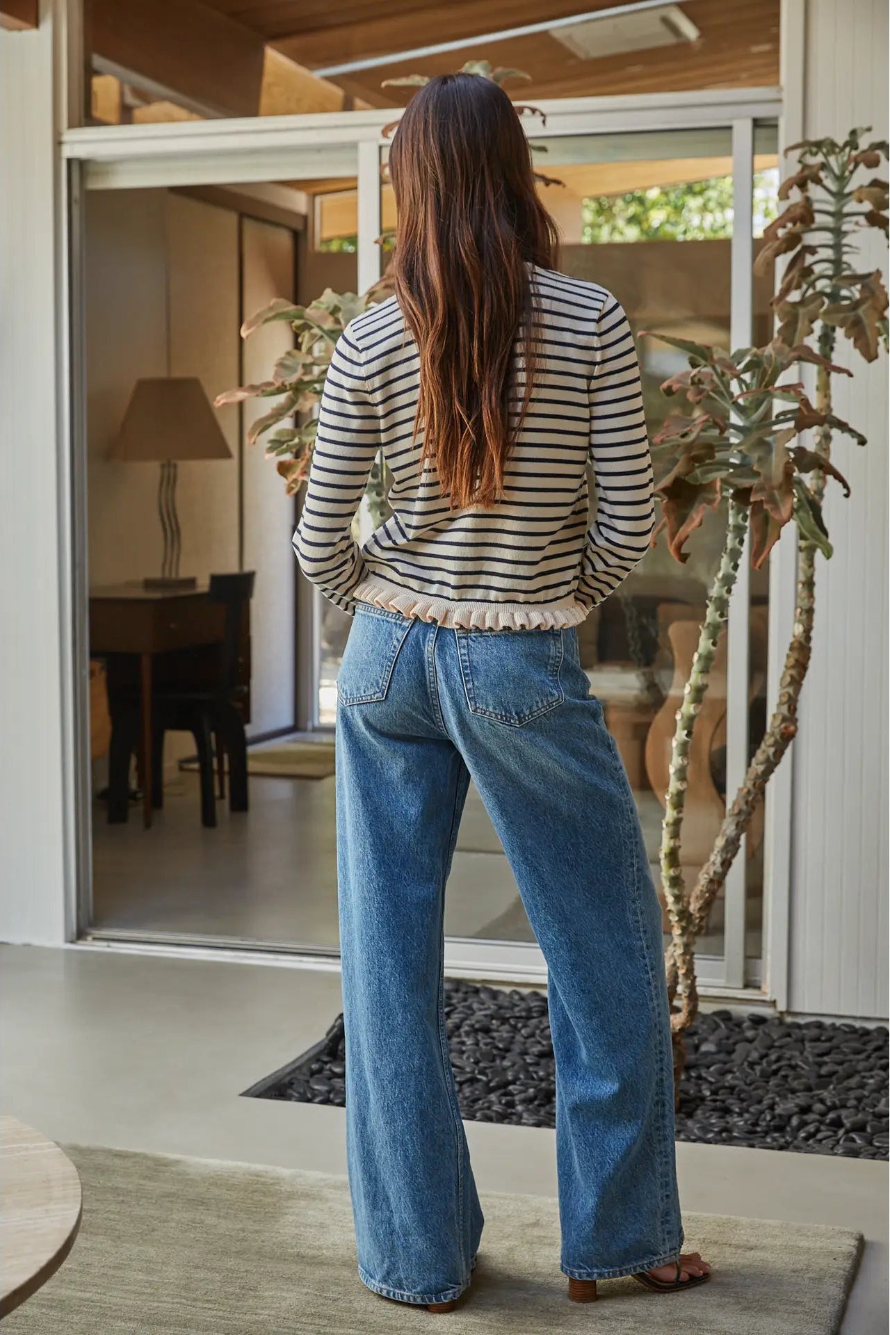 A woman viewed from behind wearing a black and white striped long-sleeve top with a ruffled hem and wide-leg blue denim jeans, standing in a modern home entryway.