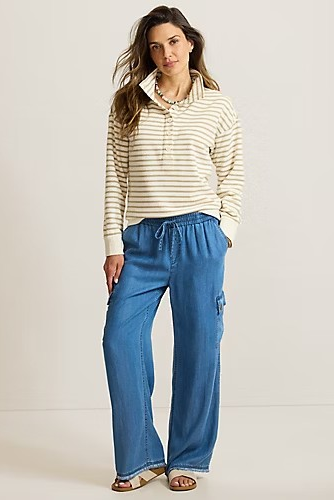 A woman modeling a cream and tan horizontally striped long-sleeve polo shirt paired with wide-leg blue jeans and beige sandals, standing against a plain light beige studio background.