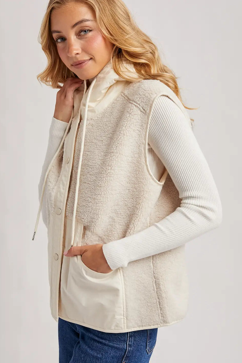 A woman wearing a cream sherpa fleece hooded vest over a white ribbed long-sleeve top and blue jeans, photographed against a light gray studio background.