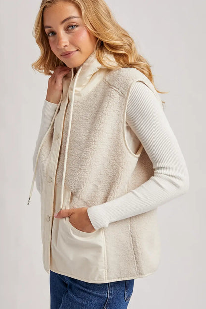 A woman wearing a cream sherpa fleece hooded vest over a white ribbed long-sleeve top and blue jeans, photographed against a light gray studio background.