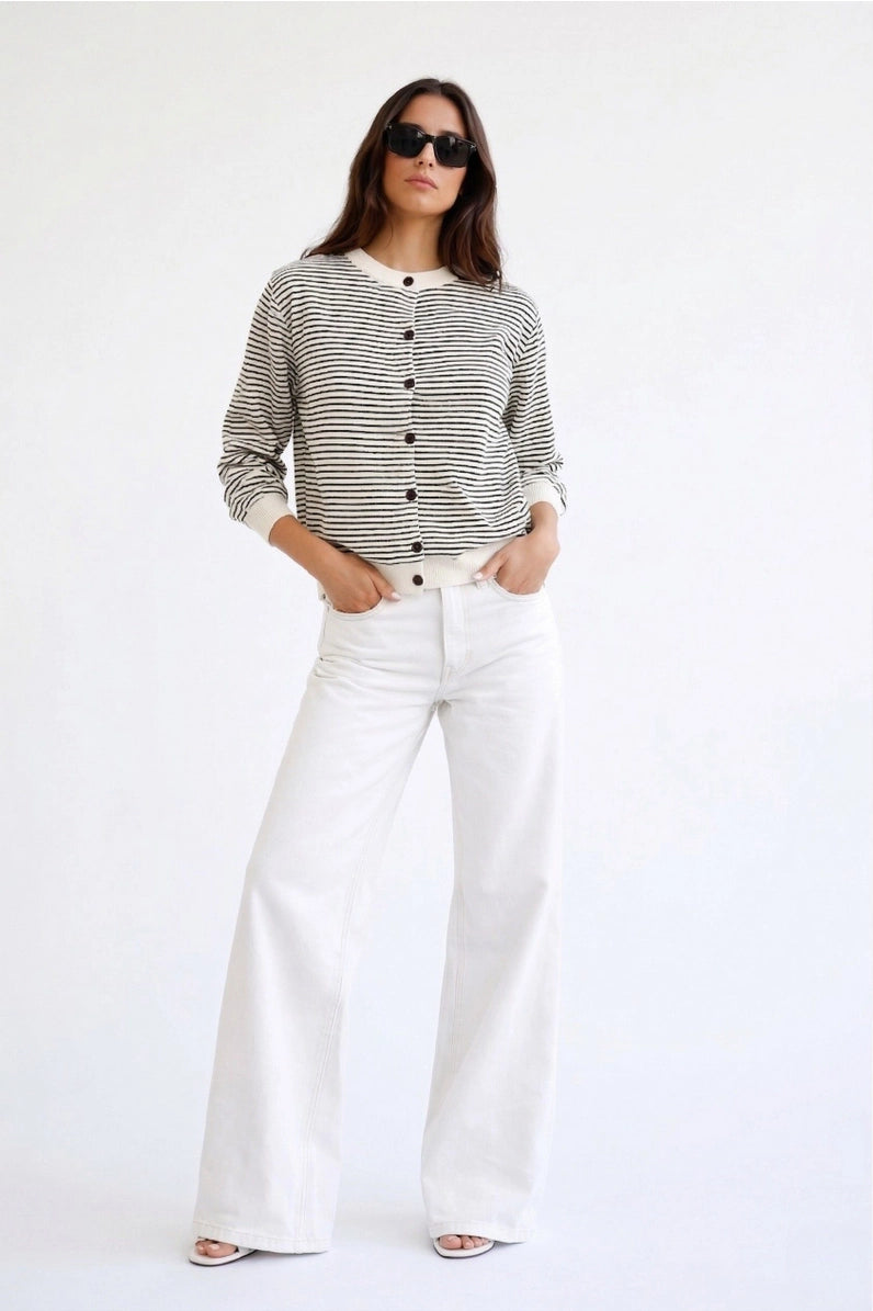 A woman modeling a black and white horizontally striped long-sleeve button-up top with white wide-leg jeans, sunglasses, and flat sandals, standing against a plain white studio background.