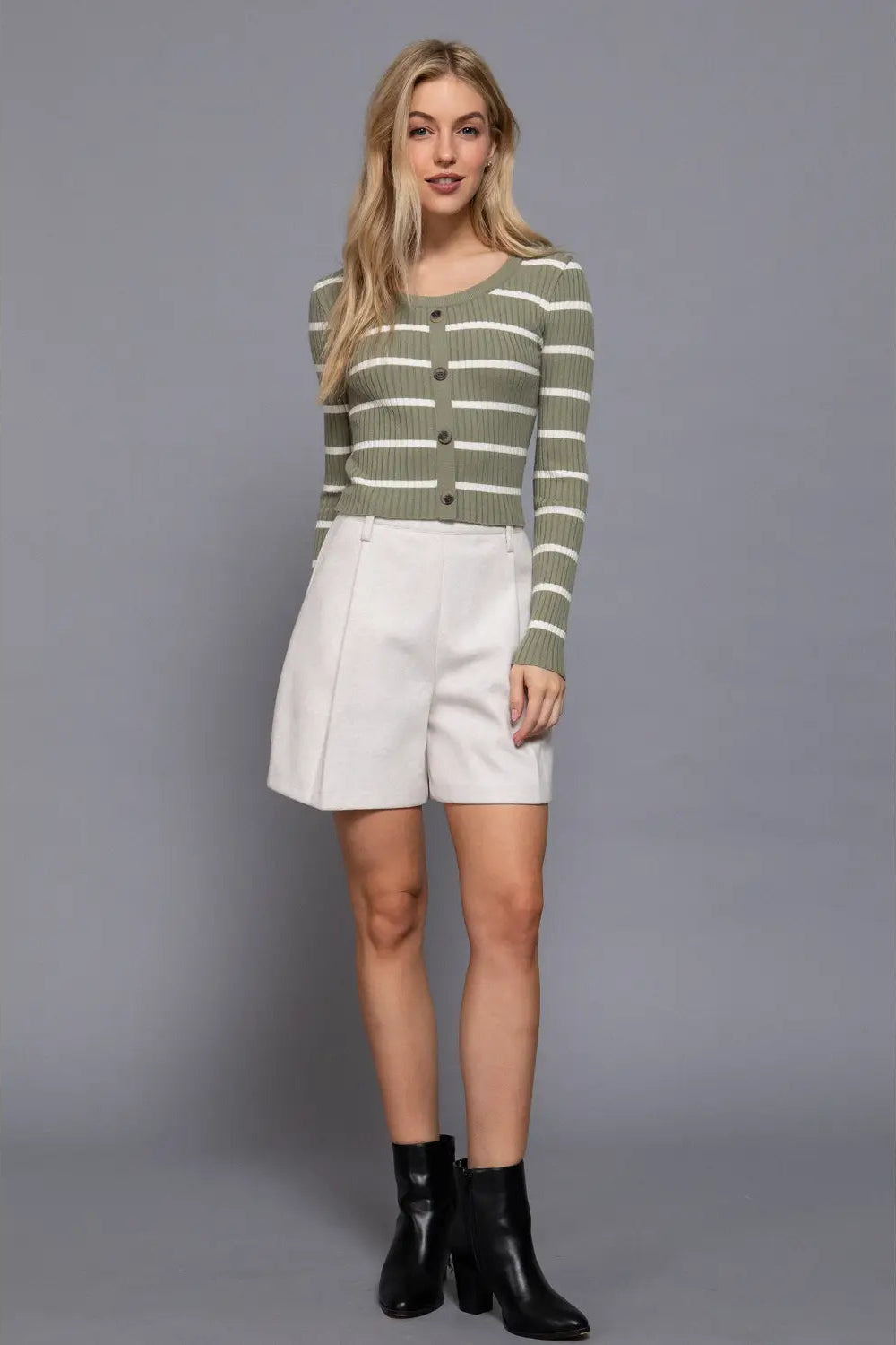 A woman modeling a green and white striped long-sleeve knit cardigan with buttons, paired with white shorts and black ankle boots, against a solid gray studio background.