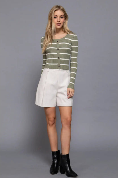 A woman modeling a green and white striped long-sleeve knit cardigan with buttons, paired with white shorts and black ankle boots, against a solid gray studio background.