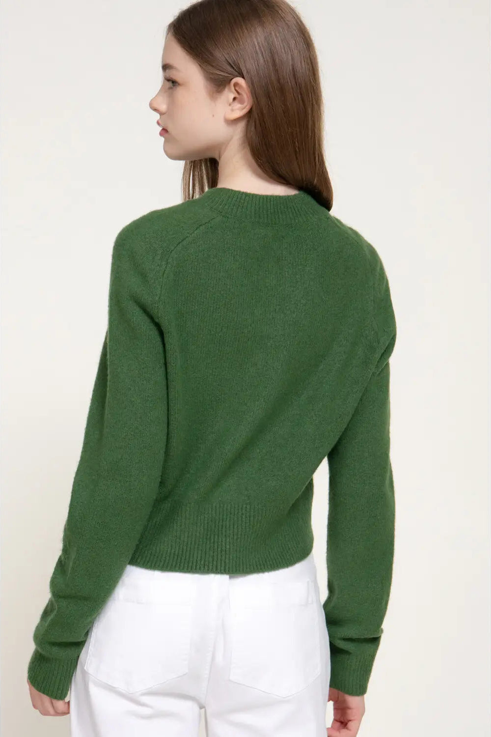 A woman shown from the back wearing a dark green knit sweater and white pants, photographed against a plain light beige studio background.