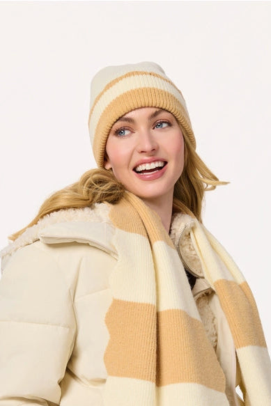 A woman wearing a cream and tan striped knit scarf and matching beanie with a cream puffer jacket and blue jeans, photographed against a plain white studio background.