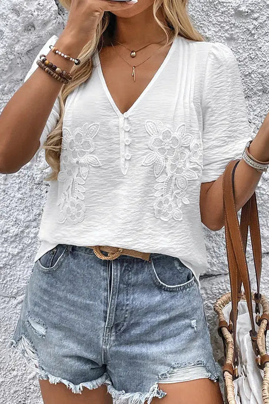 Woman wearing a white embroidered top and denim shorts against a textured wall.
