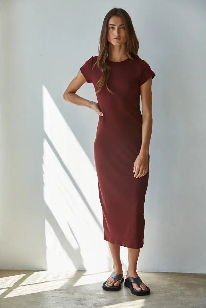 A woman modeling a short-sleeve, knee-length burgundy knit dress with black slide sandals, standing in a brightly lit room with diagonal sunlight shadows on a white wall.