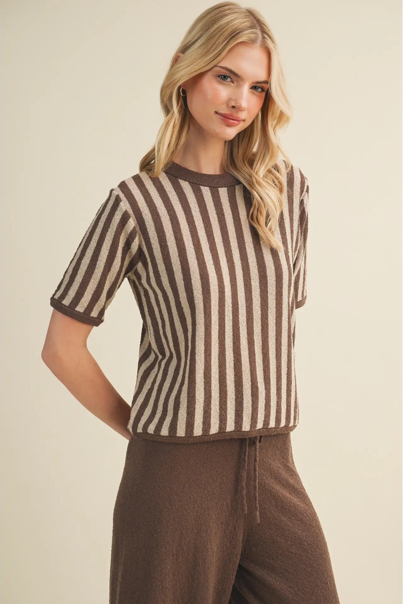 A woman modeling a short-sleeve knit top with vertical brown and cream stripes, paired with brown drawstring pants, against a plain beige studio background.