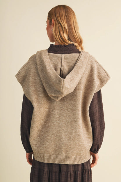 A woman shown from the back wearing a light beige knit hooded sweater vest over a dark brown long-sleeve top and dark skirt, against a plain cream background.