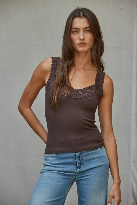 A woman modeling a dark brown sleeveless top with lace trim at the scoop neckline, paired with light blue jeans, set against a neutral gray wall.
