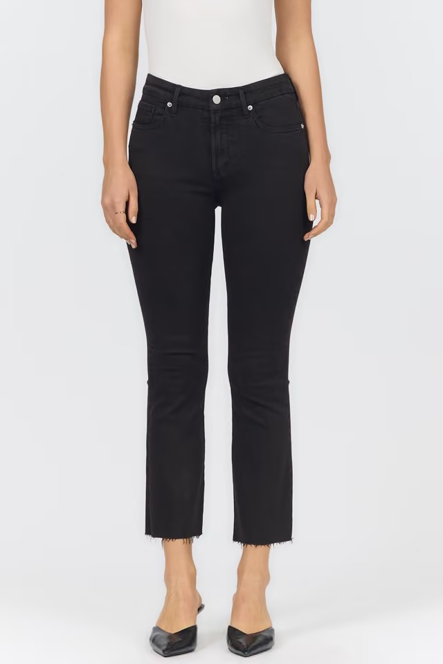 A model wearing black cropped ankle jeans with a raw hem, paired with a white top and black pointed-toe heels, shown against a plain light gray studio background.
