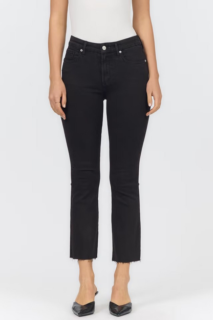 A model wearing black cropped ankle jeans with a raw hem, paired with a white top and black pointed-toe heels, shown against a plain light gray studio background.