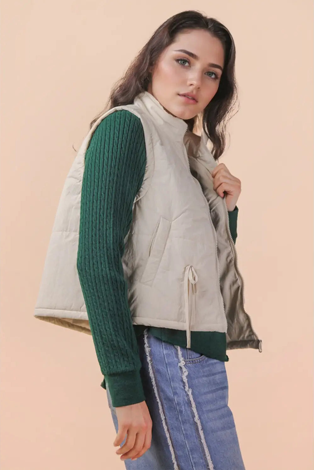 A woman wearing a light beige quilted sleeveless puffer vest over a dark green ribbed long-sleeve sweater and blue jeans, posed against a solid pale peach background.