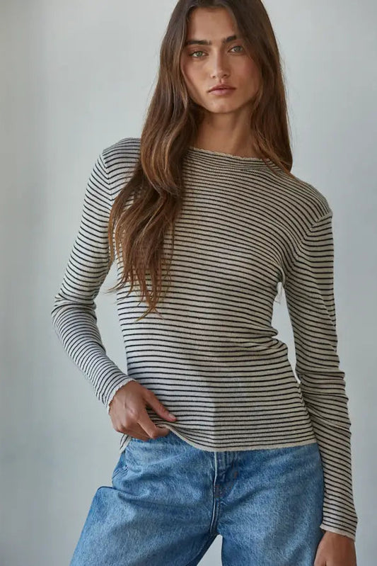 A woman wearing a fitted long-sleeve striped knit top with blue jeans, standing against a plain light gray studio background.